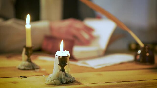 Scene from the 18th century of a man in the background using a feather quill pen to write in a journal with candles dripping with melted wax as focus in the foreground.