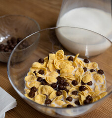 Quick cereal breakfast. Corn flakes with chocolate balls and milk in a glass bowl on a wooden table with glass jug of milk and a bowl of chocolate balls on it. Healthy food concept.