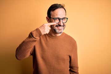 Middle age hoary man wearing brown sweater and glasses over isolated yellow background Pointing with hand finger to face and nose, smiling cheerful. Beauty concept