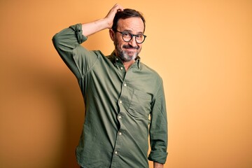 Middle age hoary man wearing casual green shirt and glasses over isolated yellow background confuse and wonder about question. Uncertain with doubt, thinking with hand on head. Pensive concept. - obrazy, fototapety, plakaty