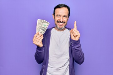 Middle age handsome man holding bunch of dollars banknotes over isolated purple background smiling with an idea or question pointing finger with happy face, number one
