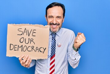 Middle age business man wearing usa tie holding banner with save our democracy message screaming proud, celebrating victory and success very excited with raised arm