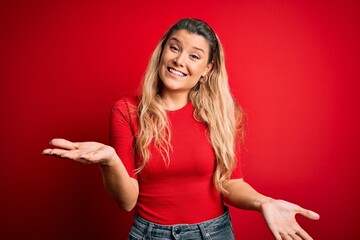 Young beautiful blonde woman wearing casual t-shirt standing over isolated red background smiling cheerful with open arms as friendly welcome, positive and confident greetings