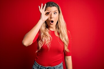 Young beautiful blonde woman wearing casual t-shirt standing over isolated red background doing ok gesture shocked with surprised face, eye looking through fingers. Unbelieving expression.