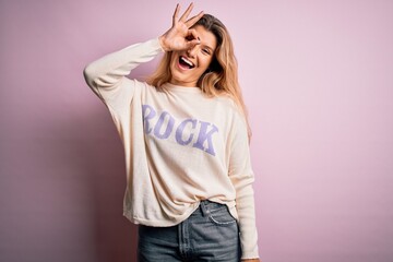 Young beautiful blonde woman wearing casual sweater standing over pink background doing ok gesture with hand smiling, eye looking through fingers with happy face.