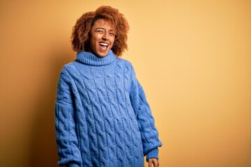 Young beautiful African American afro woman with curly hair wearing blue turtleneck sweater winking...