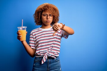 Young African American woman with curly hair on vacation wearing hat drinking orange juice with angry face, negative sign showing dislike with thumbs down, rejection concept