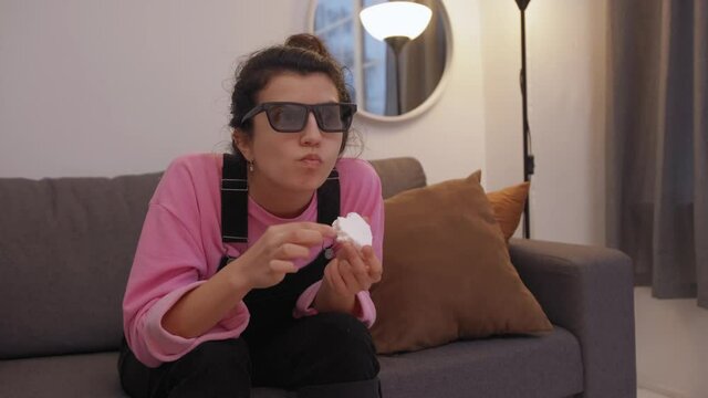 front view of woman in 3d glasses sitting on sofa and eating zefir while watching tv in living room.