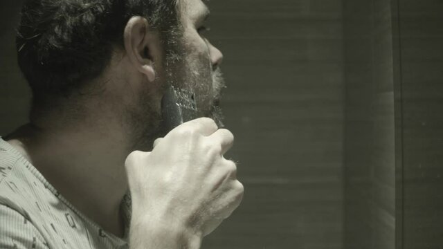 The guy shaves his beard with a trimmer in front of the bathroom mirror