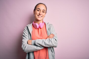 Beautiful sporty woman doing sport listening to music using headphones over pink background happy face smiling with crossed arms looking at the camera. Positive person.