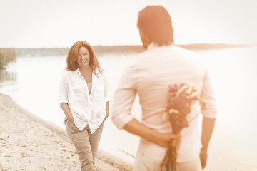 Romantic meeting of loving couple on sandy beach near sea. Focus on mature smiling woman looking at man holding bouquet of wildflowers behind his back while standing in the foreground. Toned image.