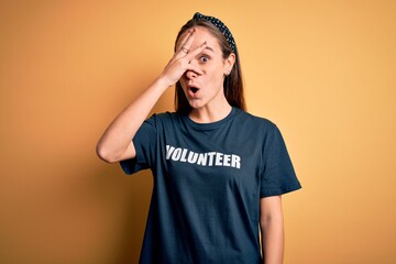 Young beautiful woman wearing volunteer t-shirt doing volunteering over yellow background peeking...