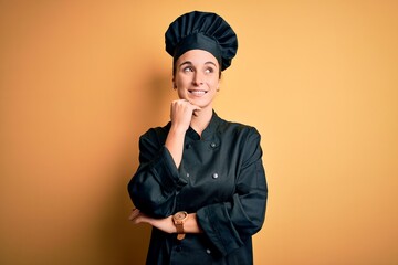 Young beautiful chef woman wearing cooker uniform and hat standing over yellow background with hand on chin thinking about question, pensive expression. Smiling and thoughtful face. Doubt concept.