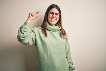 Young beautiful woman wearing casual sweater standing over isolated white background smiling and confident gesturing with hand doing small size sign with fingers looking and the camera. Measure