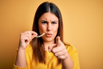 Young beautiful woman holding dental orthodontic aligner corrector over yellow background pointing with finger to the camera and to you, hand sign, positive and confident gesture from the front