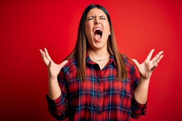Young beautiful woman wearing casual shirt over red background crazy and mad shouting and yelling...