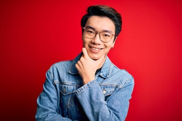 Fototapeta premium Young handsome chinese man wearing denim jacket and glasses over red background looking confident at the camera smiling with crossed arms and hand raised on chin. Thinking positive.