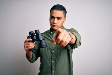 Young brazilian man looking through binoculars over isolated white background pointing with finger...