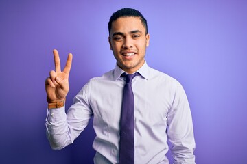 Young brazilian businessman wearing elegant tie standing over isolated purple background smiling looking to the camera showing fingers doing victory sign. Number two.