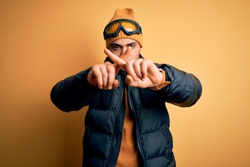 Young brazilian skier man wearing snow sportswear and ski goggles over yellow background Rejection expression crossing fingers doing negative sign