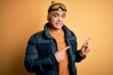 Young brazilian skier man wearing snow sportswear and ski goggles over yellow background Pointing aside worried and nervous with both hands, concerned and surprised expression