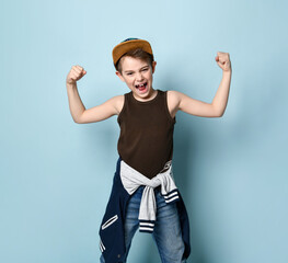 Teenage male in cap and casual stylish clothes. He clenched his fists showing muscles and screaming, posing on blue background