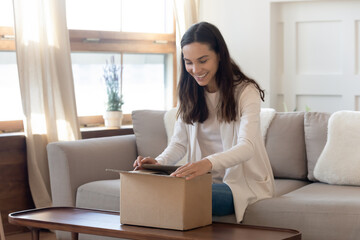 Smiling millennial girl unpack cardboard postal box with order shopping online, happy young woman...