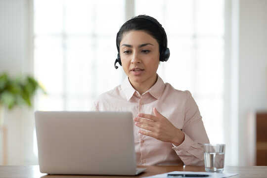 Focused successful young indian businesswoman wearing wireless headset, holding video call with partners in office. Concentrated millennial professional teacher giving online language class to client.