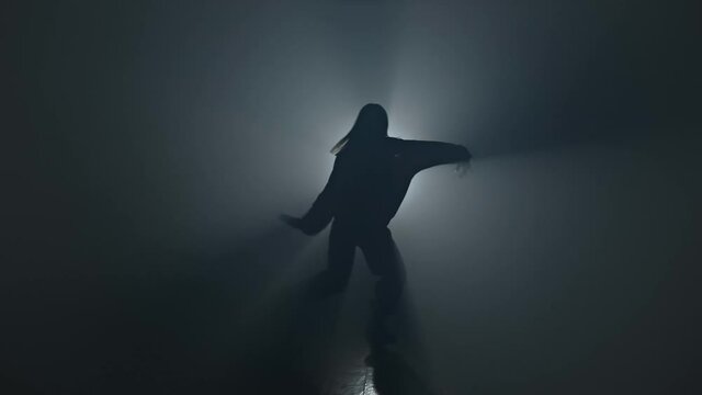 Silhouette of talented female dancer dancing hip hop in smoky studio. Young woman dancing on stage with smoke and spotlight in 4K, UHD