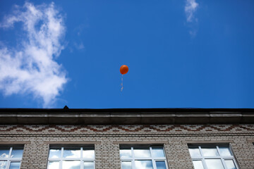 Fototapeta na wymiar Orange balloon in the blue sky above the school. End of school year. School building, school windows.