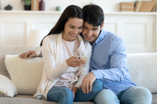 Happy millennial caucasian couple sit relax on couch at home brose wireless internet on smartphone together, smiling young man and woman rest on sofa enjoy funny video, use modern cellphone gadget