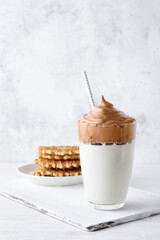 Homemade Dalgona coffee with waffles, light concrete background. Tall transparent glass with beverage and paper tube. Korean coffee cold drink with whipped instant coffee and milk. Copy space