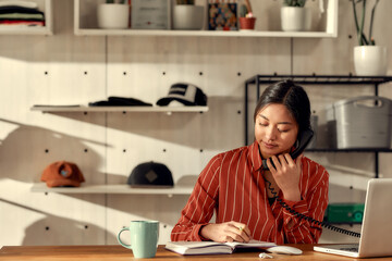 Focused on client. Portrait of young female worker receiving orders online or via telephone while sitting in the office. Woman working at custom T-shirt, clothing printing company