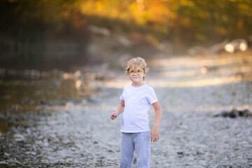 Little blonde boy playing on the riverside. Autumn in the yellow forest