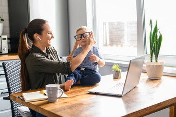 Mother works from home. Mom sits in the kitchen with laptop, kid is putting her eyeglasses on.