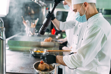 Chefs in protective masks and gloves prepare food in the kitchen of a restaurant or hotel.