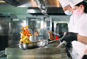 Chefs in protective masks and gloves prepare food in the kitchen of a restaurant or hotel.