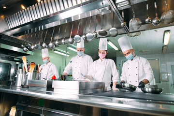 Chefs in protective masks and gloves prepare food in the kitchen of a restaurant or hotel.