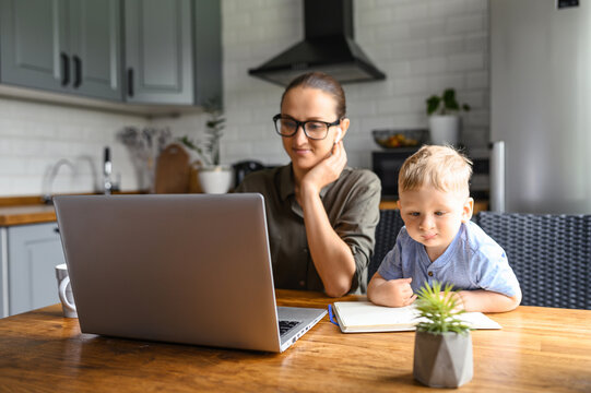 Business Mom Works From Home. Young Woman Working On A Distance With Laptop In The Kitchen, A Little Son Is Near.