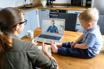 Mother work from home. Mom talks online, has video meeting, a kid sits near in the kitchen
