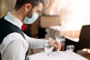 A waiter in a medical protective mask serves the table in the restaurant.