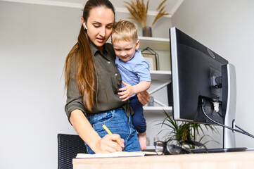 A young mother in casual wear working remotely, woman with a toddler on the arms writes notes in planner on a pc desk in home office