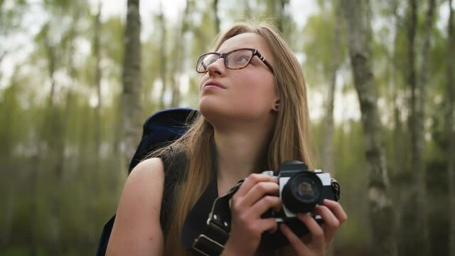 Face expression of a young caucasian woman traveler with backpack and vintage camera surrounded by trees. Portrait slow motion shot.