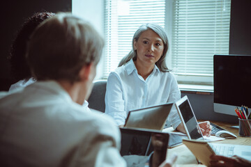 Office staff brainstorming. Elderly Asian woman listens attentively looking at her colleagues while sitting at negotiating table during a business meeting.