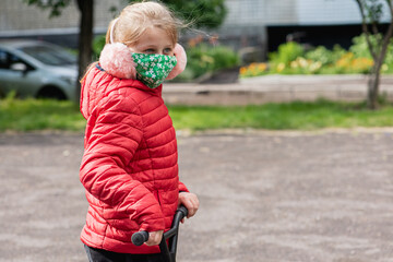 Fototapeta premium little blonde, pretty girl sitting on the playground, in a medical mask, with a scooter, during quarantine, due to a pandemic, isolation 