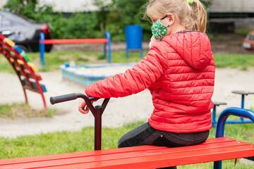 Obraz premium little blonde, pretty girl sitting on the playground, in a medical mask, with a scooter, during quarantine, due to a pandemic, isolation 