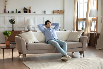 Relaxed young man sit rest on couch in modern design living room daydreaming or sleeping, calm millennial male relax on sofa at home, breathe fresh air, relieve negative emotion, stress free concept
