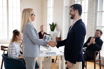 Smiling middle aged female team leader shaking hands with young motivated male colleague in suit, praising promoting while teammates applauding. Happy coworkers celebrating business achievement.