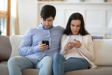 Obraz premium Young caucasian couple sit relax on couch at home busy with smartphone devices, spend weekend not talking, millennial man and woman browse internet use cellphones, gadget addiction concept