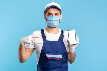 Workman in overalls, mask and gloves pointing mobile phone with mock up blank display, advertising area for online delivery order app, house repair maintenance services. indoor shot isolated on blue